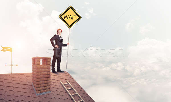 Stock photo: Young businessman on house brick roof holding yellow signboard. 