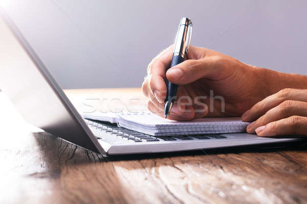 Stock photo: Person's Hand Writing On Spiral Book Over Laptop