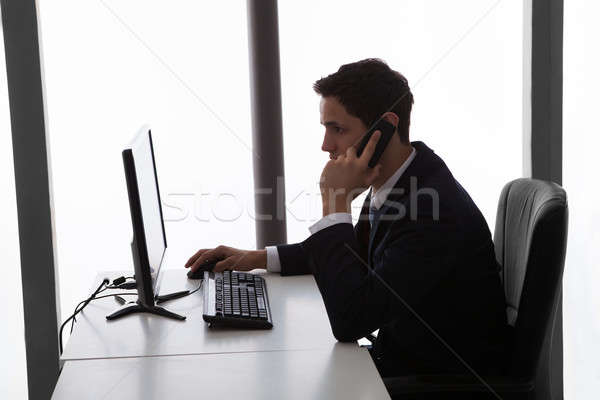 Businessman Answering Mobilephone While Using Computer At Office Stock photo © AndreyPopov