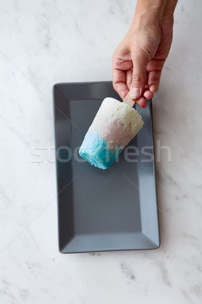A girl's hand holds a multicolored ice cream on a stick in a black plate on a gray marble background Stock photo © artjazz