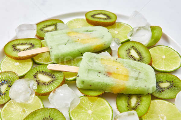 Stock photo: Close-up of homemade lime kiwi icy popsicle with a piece of peach in a plate with ice cubes and piec