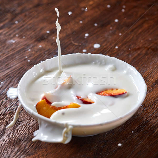 Pieces of ripe fresh peach fell in a ceramic bowl with a splash of yoghurt on a wooden table. Stock photo © artjazz