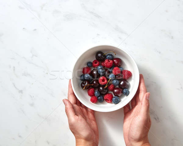 Natural organic fruits, berries in a plate on a gray stone background. Women's hands hold white bowl Stock photo © artjazz