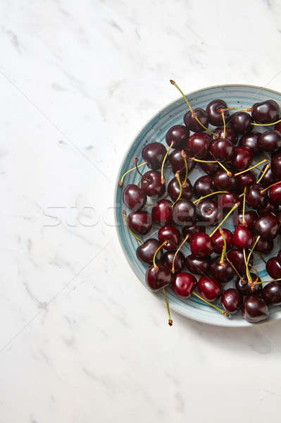 A pile of ripe sweet dark red cherries in a blue ceramic plate on a gray stone background. Flat lay, Stock photo © artjazz