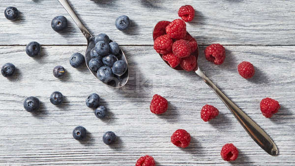 Two spoons with red juicy ripe berries - raspberry and blueberry and around them on a gray wooden ba Stock photo © artjazz