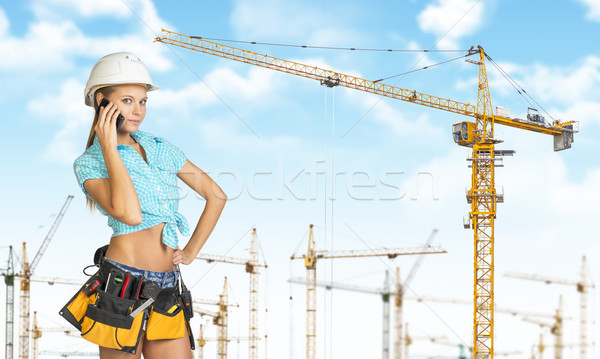 Woman in helmet and tool belt, talking on phone. Tower cranes as backdrop Stock photo © cherezoff