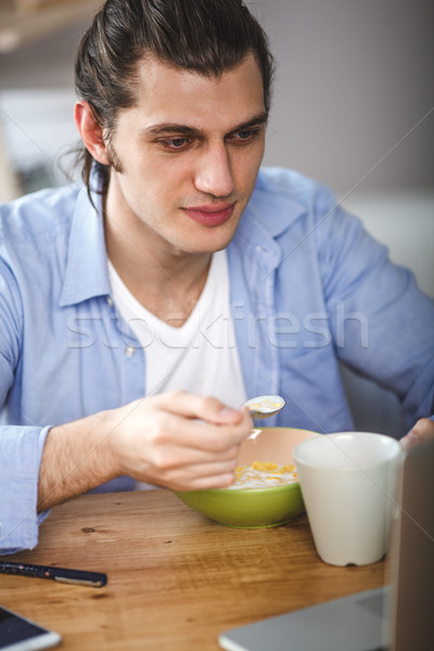 Young man eating corn flakes with milk and and looking to the laptop screen Stock photo © chesterf