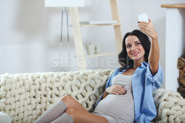 Stock photo: smiling pregnant woman taking a self-portrait with her smartphone sitting on couch