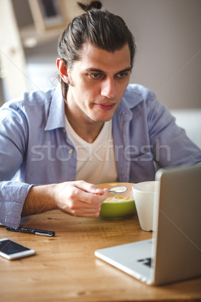 Young man eating corn flakes with milk and and looking to the laptop screen Stock photo © chesterf