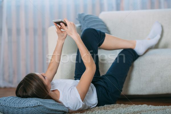 Stock photo: Young woman checking her smart phone lying on carpet