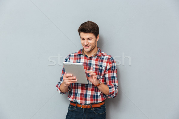 Happy young man in plaid shirt standing and using tablet Stock photo © deandrobot