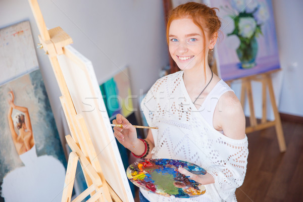Cheerful woman artist standing and painting picture in workshop Stock photo © deandrobot