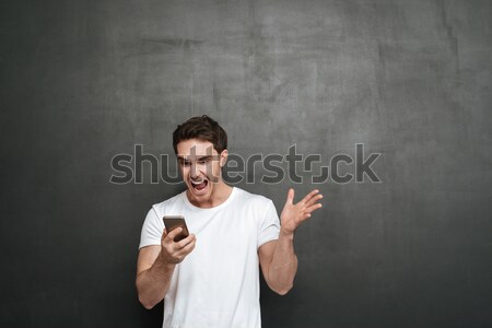 Satisfied smiling man in white shirt holding smartphone and gest Stock photo © deandrobot