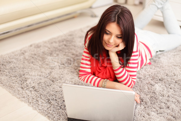 Portrait of dreamy smiling young woman using laptop while lying on floor at home Stock photo © deandrobot