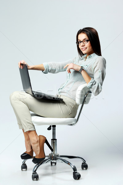 Young smiling businesswoman sitting on the office chair with laptop on gray background Stock photo © deandrobot