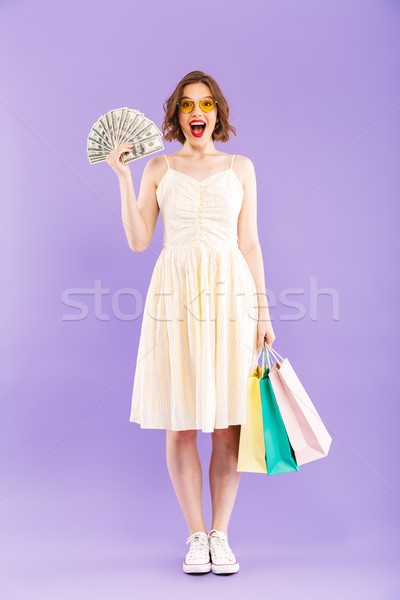 Stock photo: Excited happy woman isolated over purple wall background holding shopping bags and money.