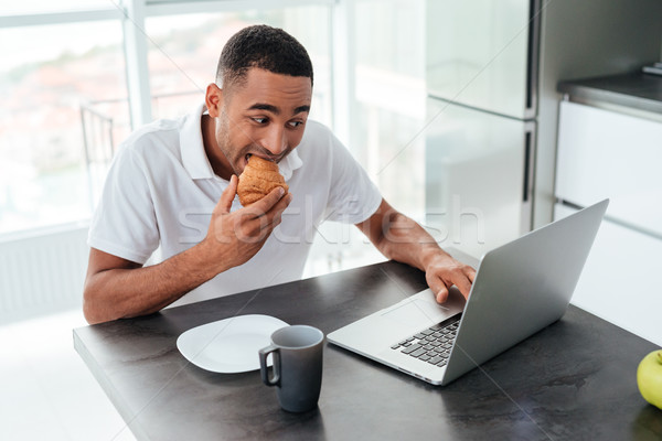 Handsome african man having breakfast and using laptop on kitchen Stock photo © deandrobot