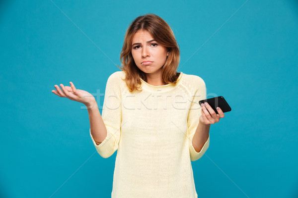 Stock photo: Confused woman in sweater holding smartphone and shrugs her shoulder