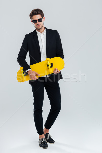 Stock photo: Young handsome businessman in suit and suglasses holding yellow skateboard