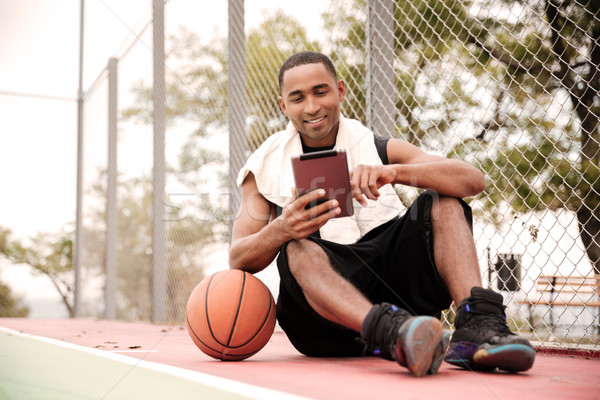 Happy african basketball player sitting in park and using tablet Stock photo © deandrobot