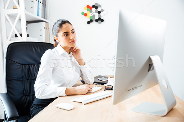 Beautiful concentrated businesswoman making notes and looking at computer screen Stock photo © deandrobot