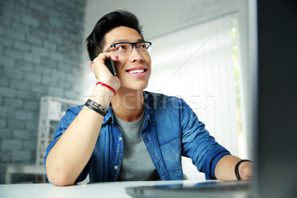 Stock photo: Young happy asian man talking on the phone at his workplace in office