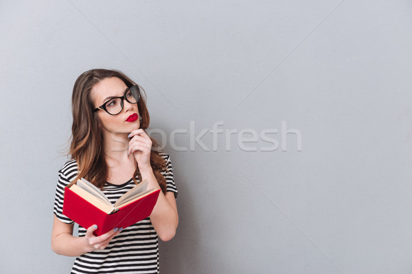 Thinking young lady standing over grey wall reading book Stock photo © deandrobot