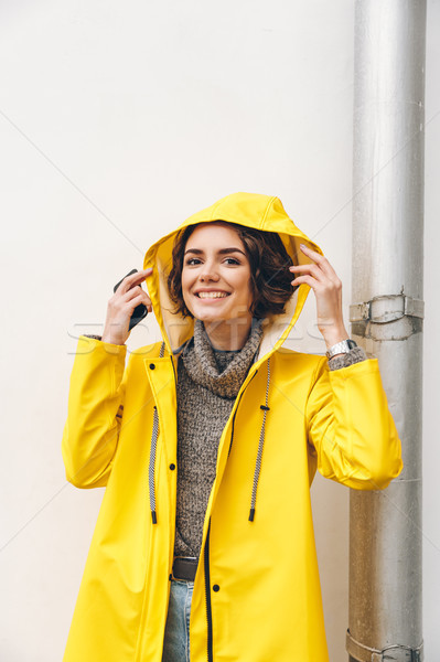 Smiling adult girl in yellow coat putting hood on head posing on Stock photo © deandrobot
