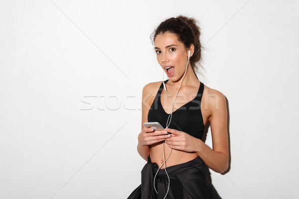 Surprised curly brunette fitness woman listening music and holding smartphone Stock photo © deandrobot