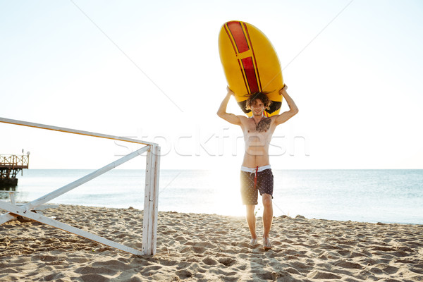Handsome surfer carrying his surfboard across beach in evening sunlight Stock photo © deandrobot