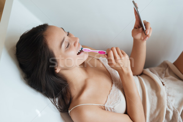 Cheerful woman bushing teeth and using mobile phone in bathtub Stock photo © deandrobot