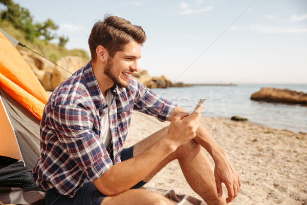 Smiling young man tourist using smartphone sitting in touristic tent Stock photo © deandrobot