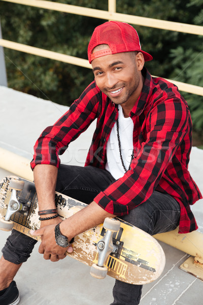 Cheerful dark skinned guy wearing cap sitting with his skateboard Stock photo © deandrobot