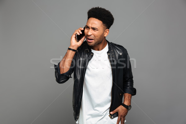 Young unhappy african guy in leather jacket speaking on mobile p Stock photo © deandrobot