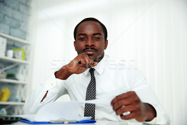 Pensive african man sitting at the table and signing document in office Stock photo © deandrobot