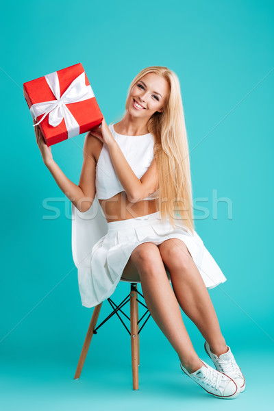 Cheerful blonde woman showing gift box and sitting on chair Stock photo © deandrobot