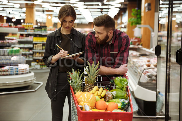 Concentrated loving couple in supermarket looking at notebook. Stock photo © deandrobot