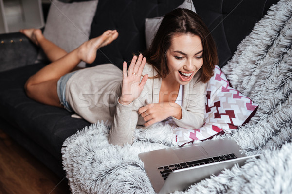 Smiling girl waving hand while lying on couch with laptop Stock photo © deandrobot