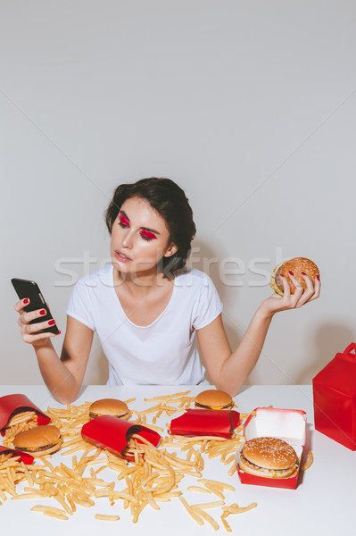 Pretty young woman with hamburger sitting and using mobile phone Stock photo © deandrobot