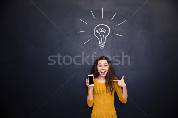 Happy woman pointing on smartphone blank screen over chalkboard background Stock photo © deandrobot