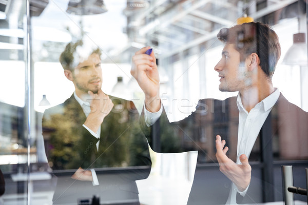 Two businessmen talking and writing on glass board in office Stock photo © deandrobot
