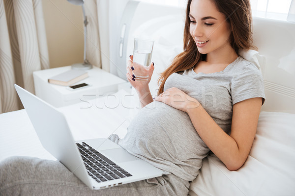 Сток-фото: улыбаясь · беременная · женщина · используя · ноутбук · питьевая · вода · кровать · беременна