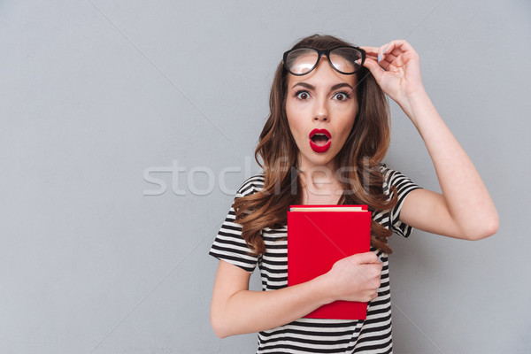 Shocked young lady standing over grey wall holding book. Stock photo © deandrobot