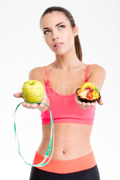 Thoughtful unsure fitness girl holding apple, measuring tape and cake Stock photo © deandrobot
