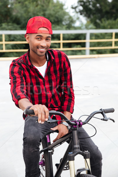 Cheerful dark skinned man wearing cap sitting on his bicycle Stock photo © deandrobot