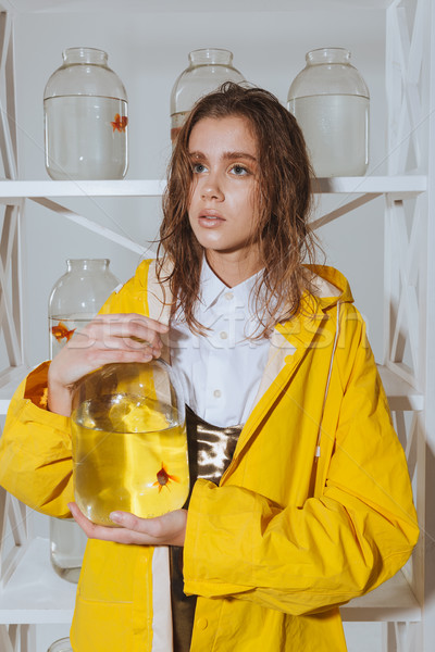 Stock photo: Pensive woman in yellow raincoat holding jar with gold fish