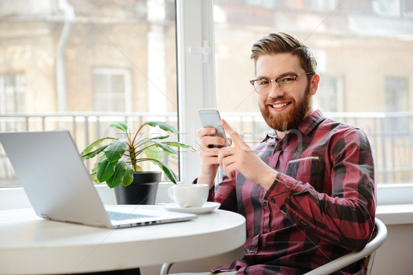 Happy bearded young man using mobile phone and laptop computer. Stock photo © deandrobot