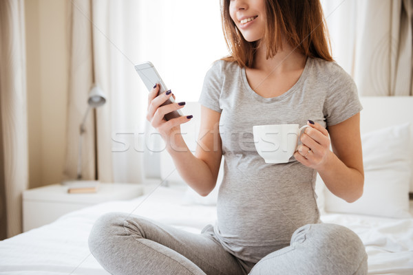 Stock photo: Happy pretty pregnant young woman drinking tea and using cell phone on bed at home