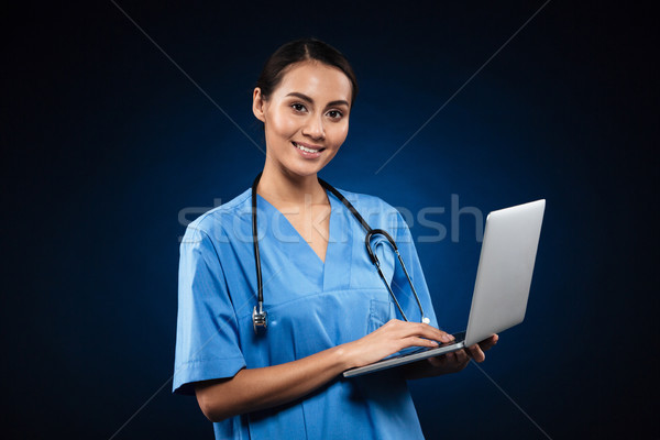 Cheerful lady in medical uniform using laptop and looking camera Stock photo © deandrobot