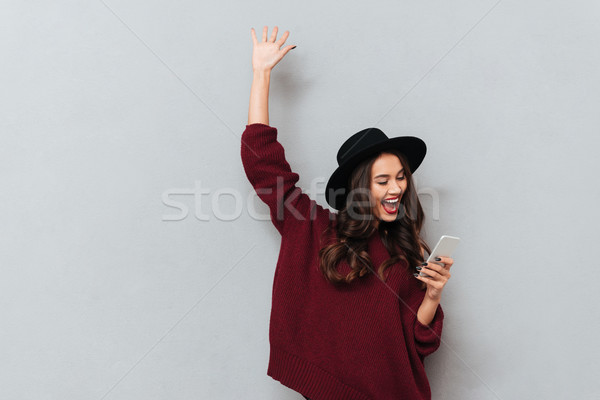 Brunette woman in sweater and hat using smartphone and rejoice Stock photo © deandrobot